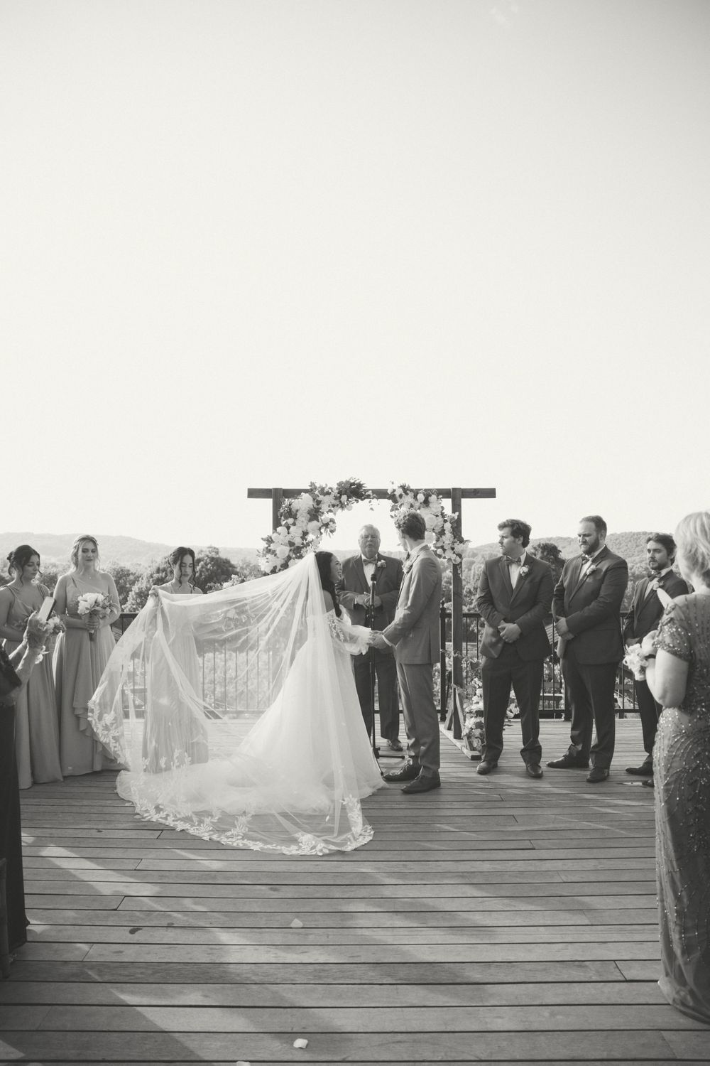 Black and white photo of wedding ceremony on rustic wooden deck with floral arch.