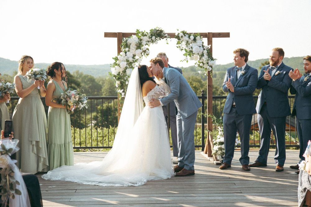 Wedding ceremony on deck with floral arch and bridal party in blue and sage green attire.