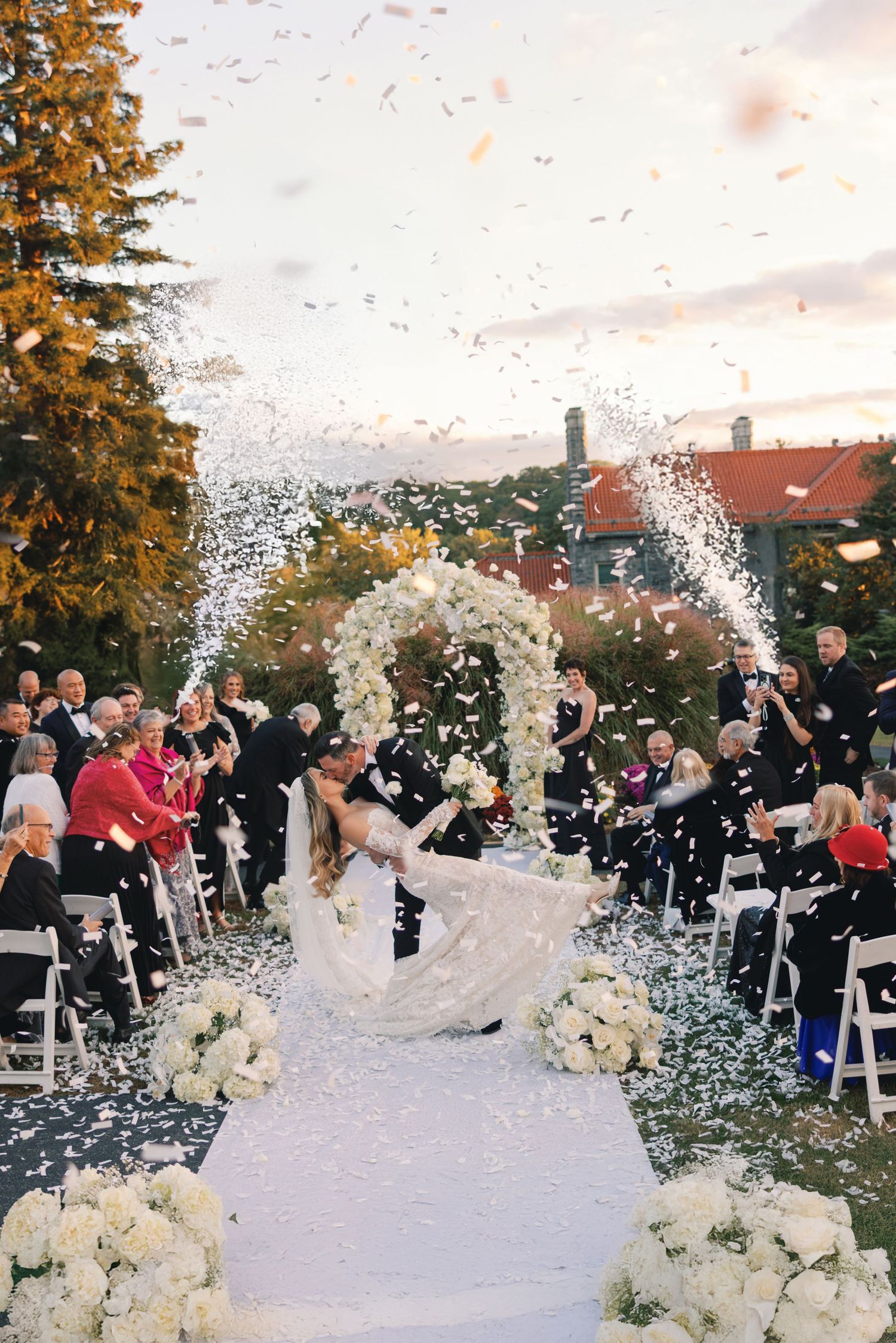 Bride and groom kissing at the altar as white confetti rains down during an elegant outdoor ceremony.