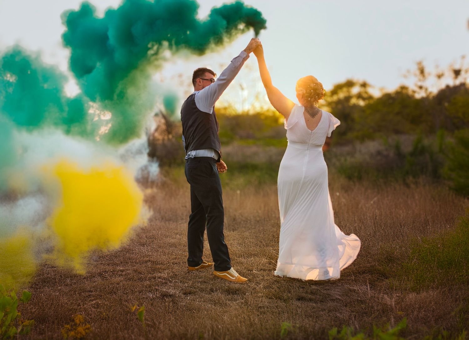 Newlyweds dance with colorful smoke bombs creating teal and yellow effects at sunset.