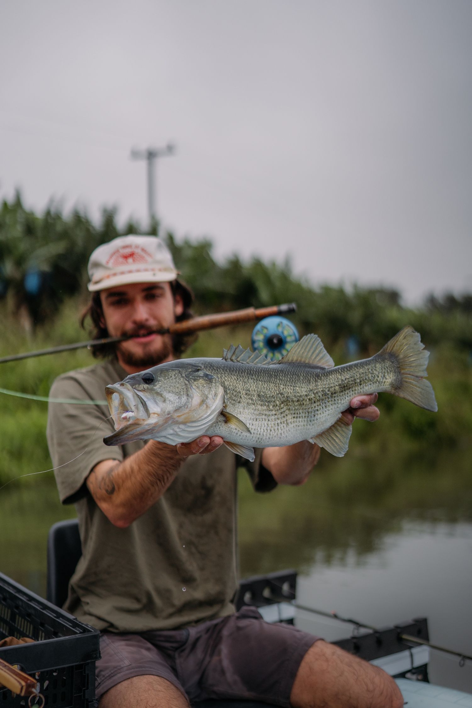 Fisherman holding a large largemouth bass caught on a fly rod while sitting in a boat on a calm waterway.