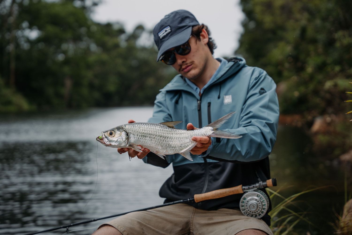 Angler in a blue jacket holds a freshly caught fish by a tranquil lake on an overcast day.
