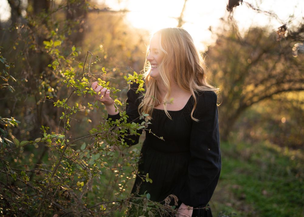 Person in black clothing near pink flowers at sunset with sunlight creating a warm backlit glow through trees.