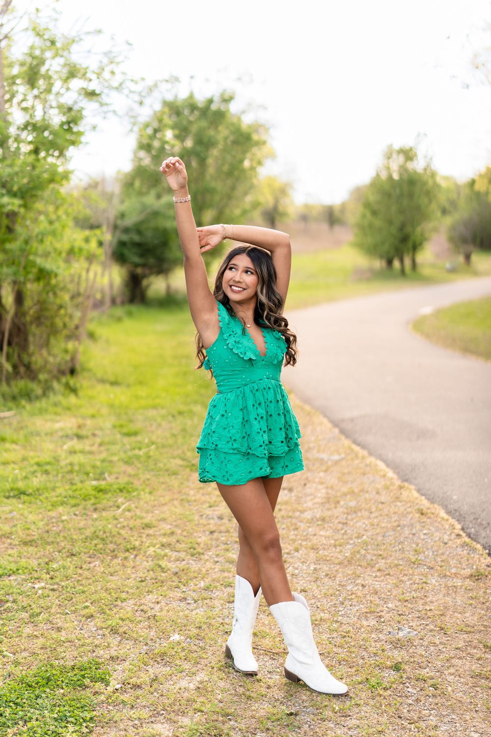 Young woman in green romper and white cowboy boots poses with arm raised on a scenic outdoor path.