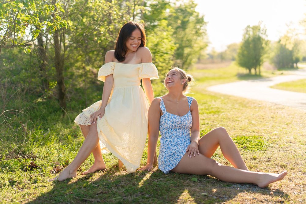 Two young women enjoying a sunny day outdoors, one in a yellow dress standing, one in floral romper sitting on grass.