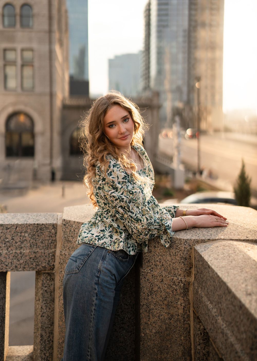 Woman in floral print blouse and jeans poses at sunset against urban architecture with golden light.