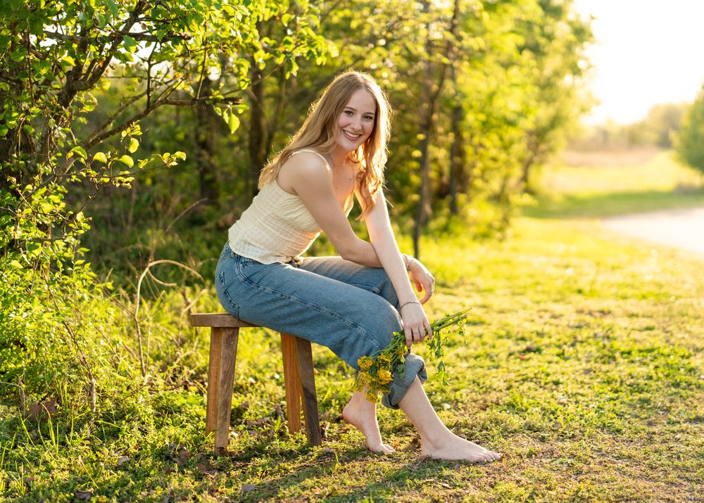 Smiling young woman sitting on a wooden bench outdoors in golden sunlight, wearing jeans and a yellow top.