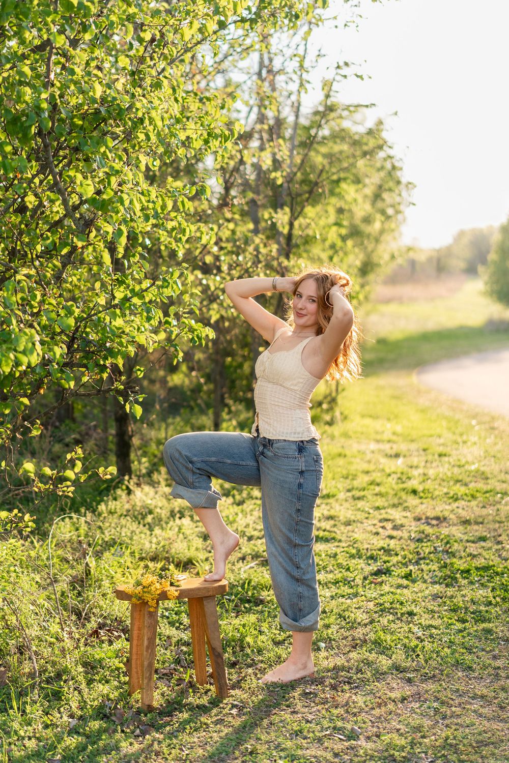 Young woman in jeans and tank top poses barefoot outdoors, resting foot on wooden stool near sunlit trees.