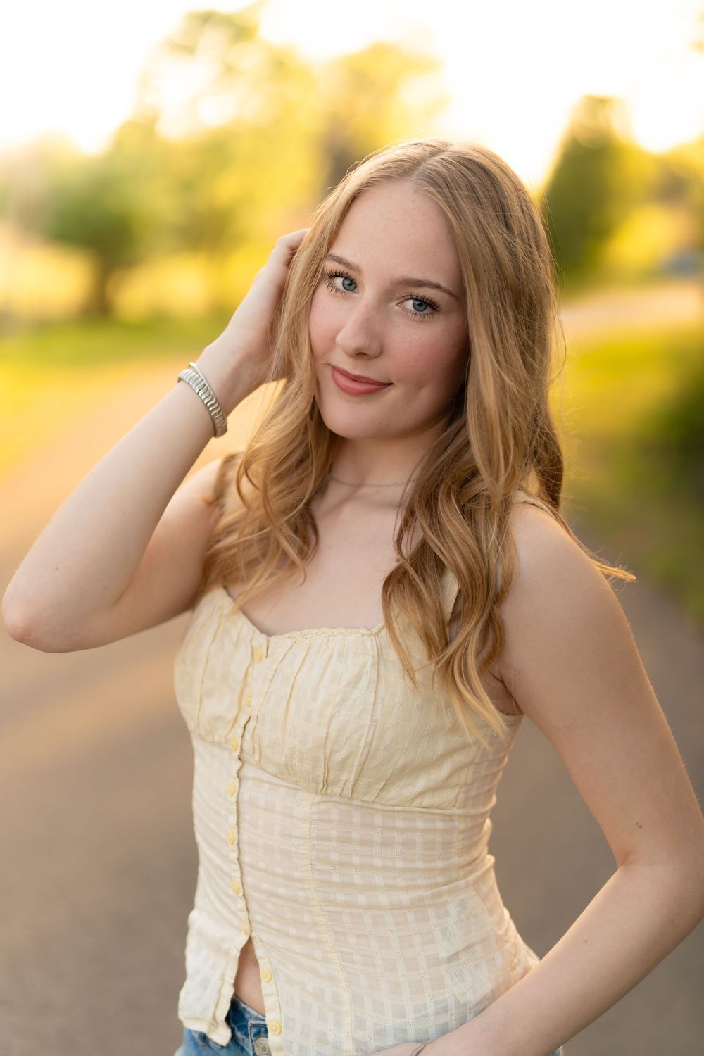 Young woman with long blonde hair in yellow top poses outdoors in golden hour sunlight.