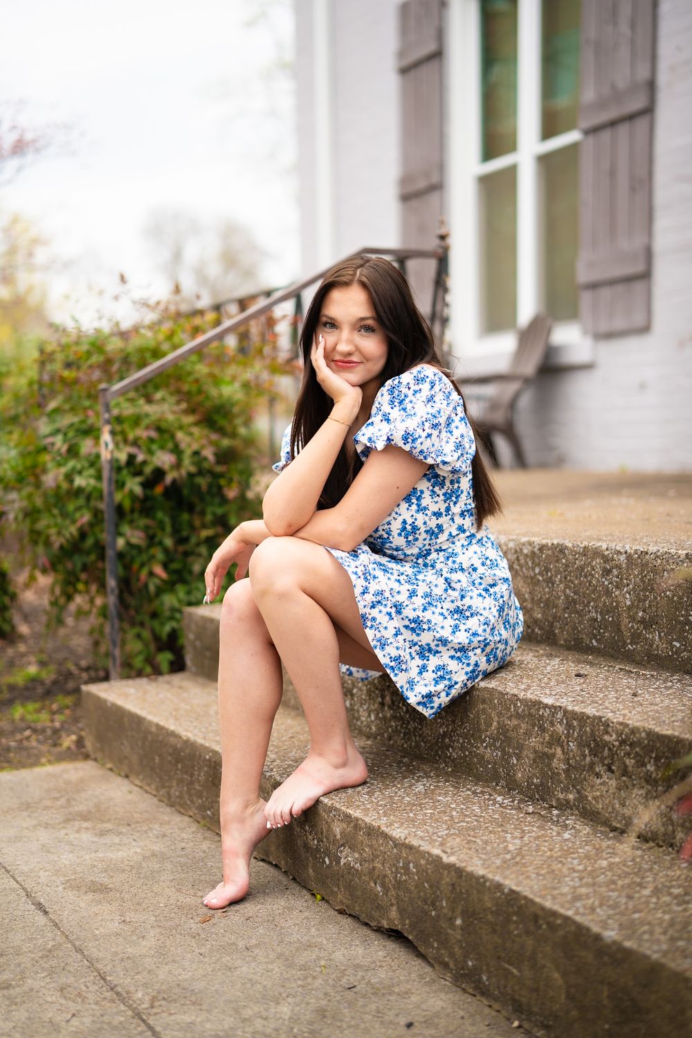 Someone in a blue and white floral dress sits barefoot on concrete steps outside a building with shutters.
