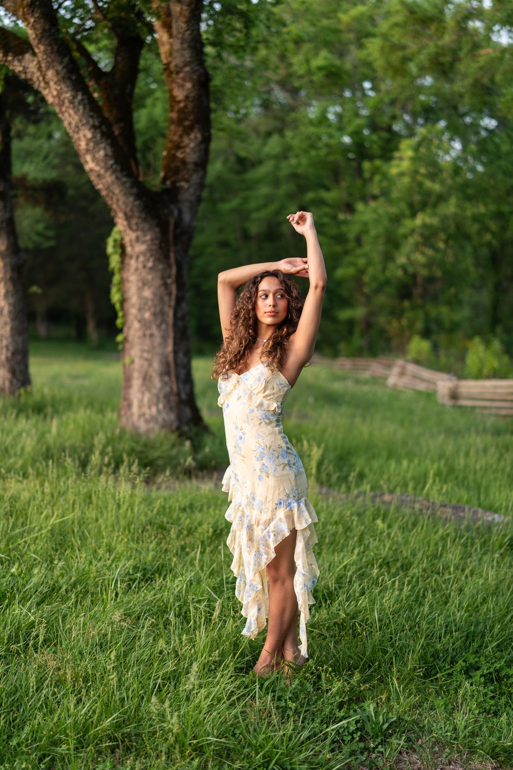 A young woman in a white floral ruffled dress poses gracefully in a lush green park with arms raised.