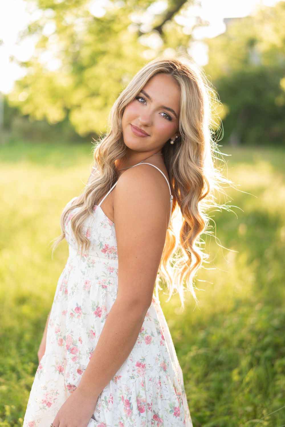 Young woman in floral sundress poses outdoors in golden sunlight with long blonde wavy hair and a warm smile.