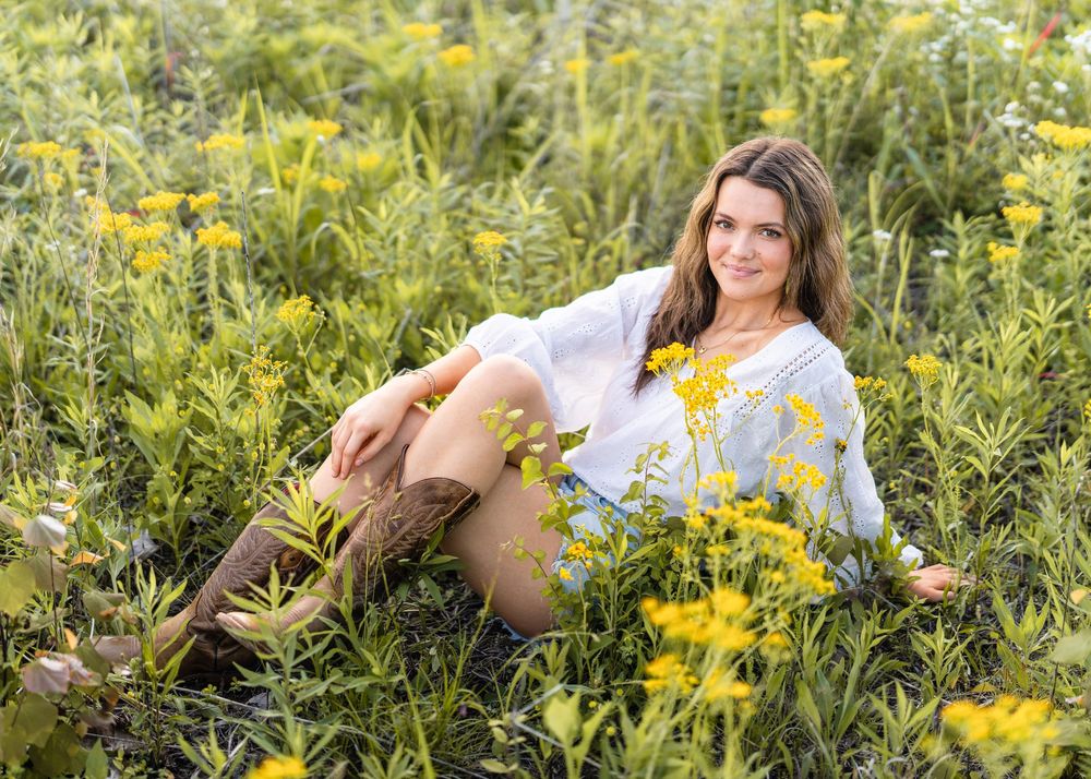 A reclining person in cowboy boots and a white floral dress poses among blooming yellow wildflowers in a summer meadow.