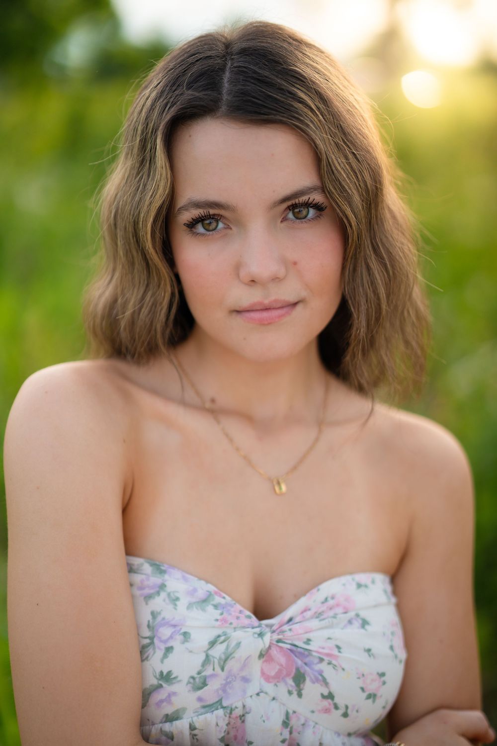 Portrait photograph outdoors with soft sunset lighting showing wavy hair and floral strapless top against blurred nature background.
