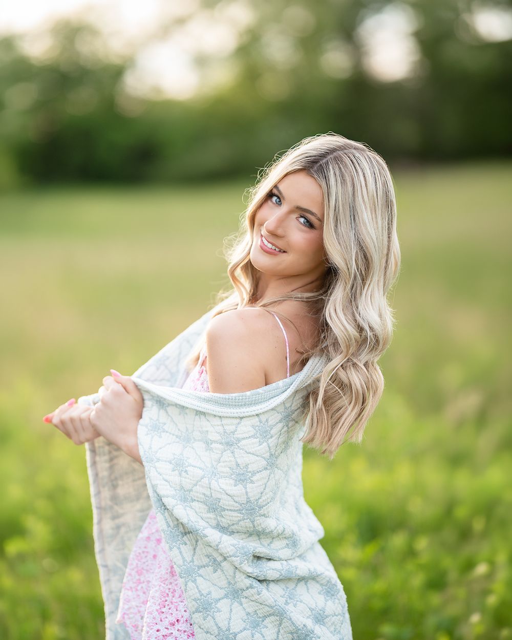 Young blonde woman smiling over shoulder in green field, wearing pink dress and holding light blue patterned wrap.