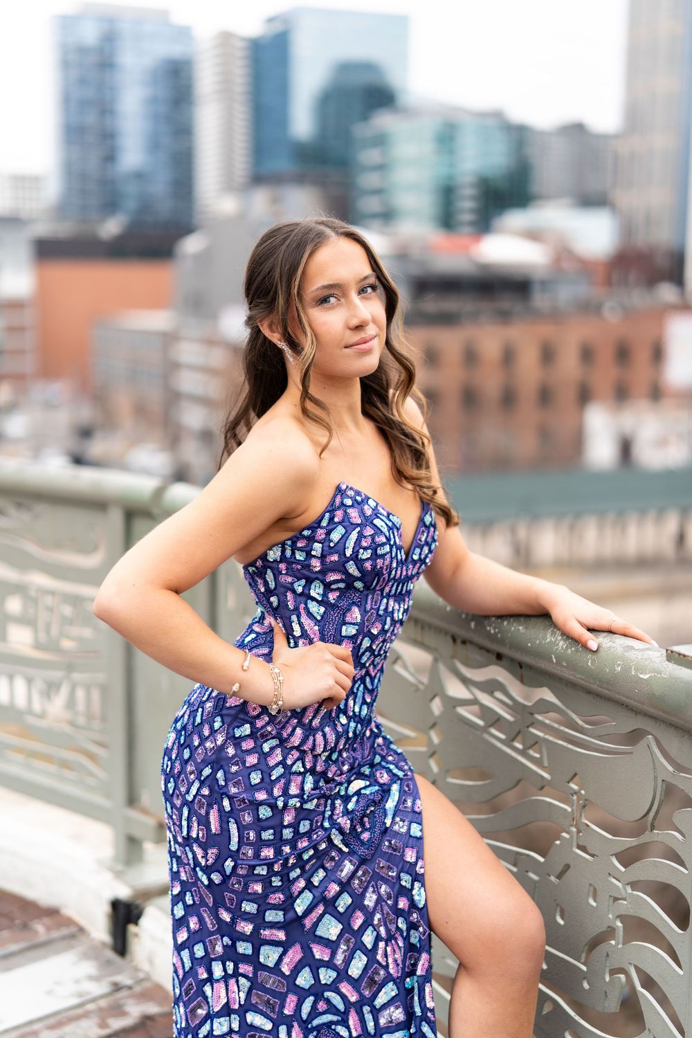 Young woman in blue patterned strapless gown poses on urban rooftop railing with city skyline backdrop.
