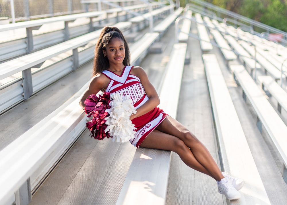 A cheerleader in red and white uniform poses confidently on bleachers with pom poms at an outdoor stadium.