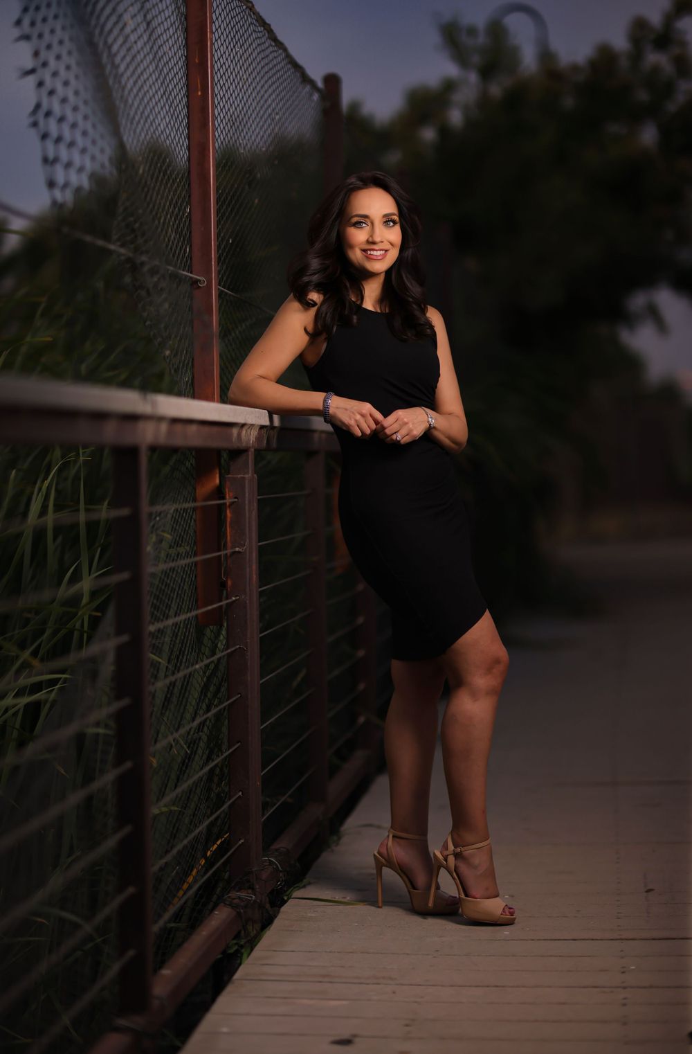 Woman in black dress poses against metal railing during evening photoshoot.