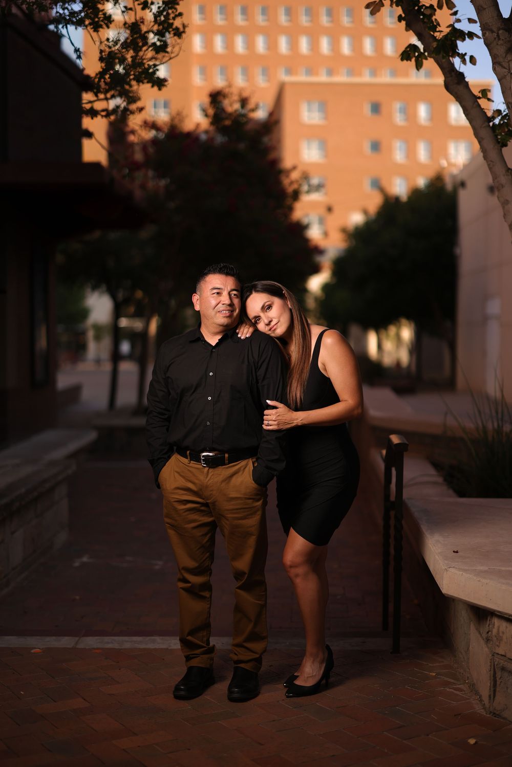 A couple shares a romantic moment at sunset against an orange brick building backdrop.