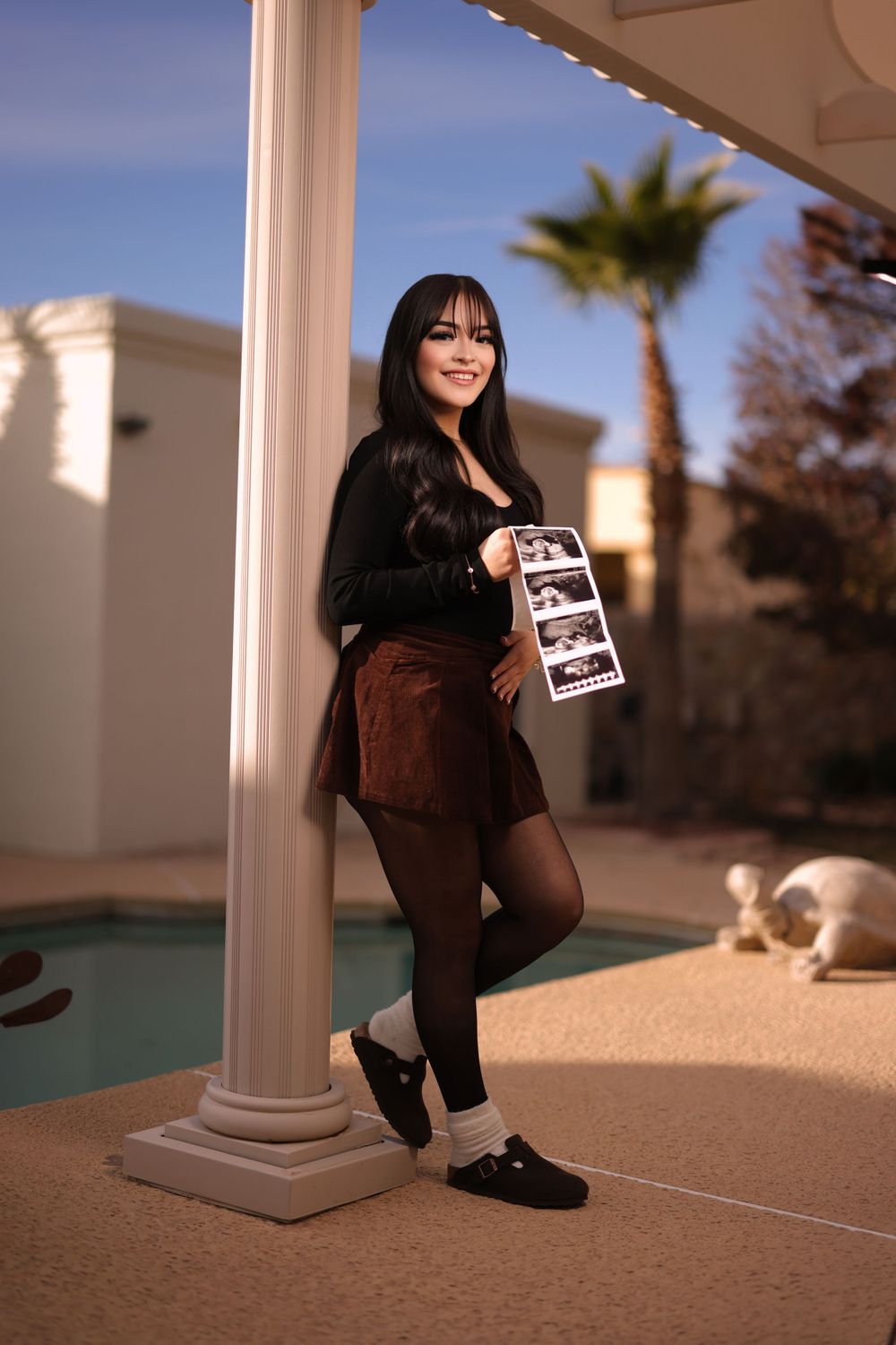 A casual portrait by a poolside pillar holding ultrasound photos.