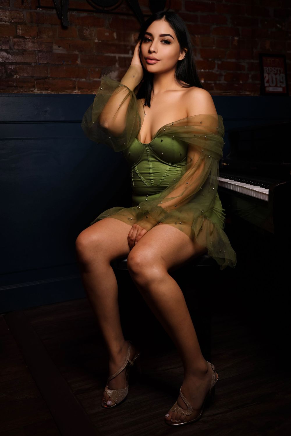 A woman in an olive green tulle dress poses seductively against a brick wall background in moody lighting.