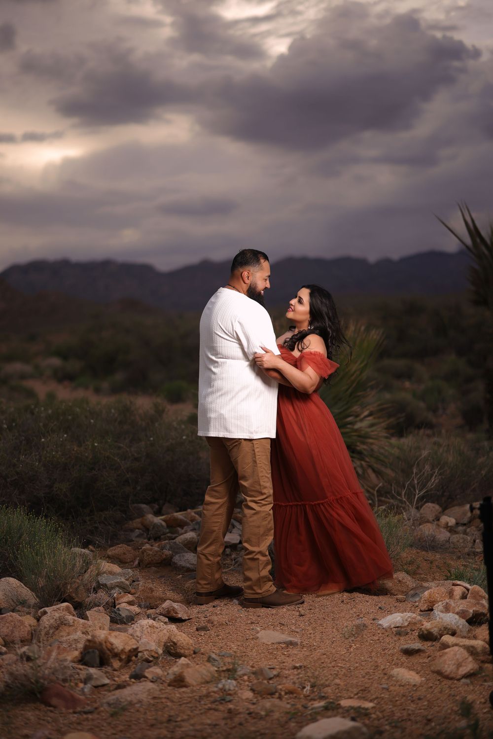 A couple shares an intimate moment on rocky desert terrain against a dramatic cloudy sky at sunset.