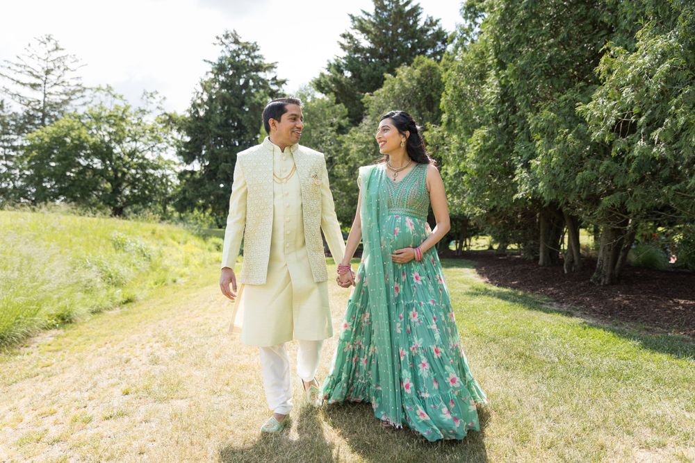 A couple holds hands while walking outdoors, wearing coordinated Indian formal wear in cream and turquoise.
