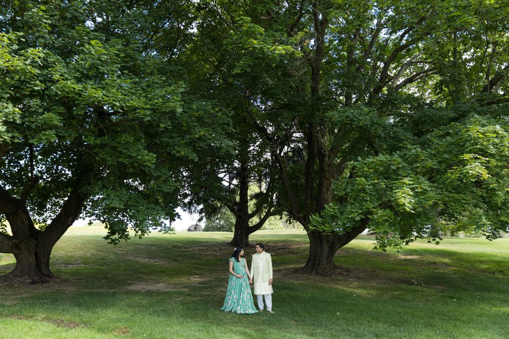 A couple in traditional dress standing beneath large trees in a peaceful park setting.