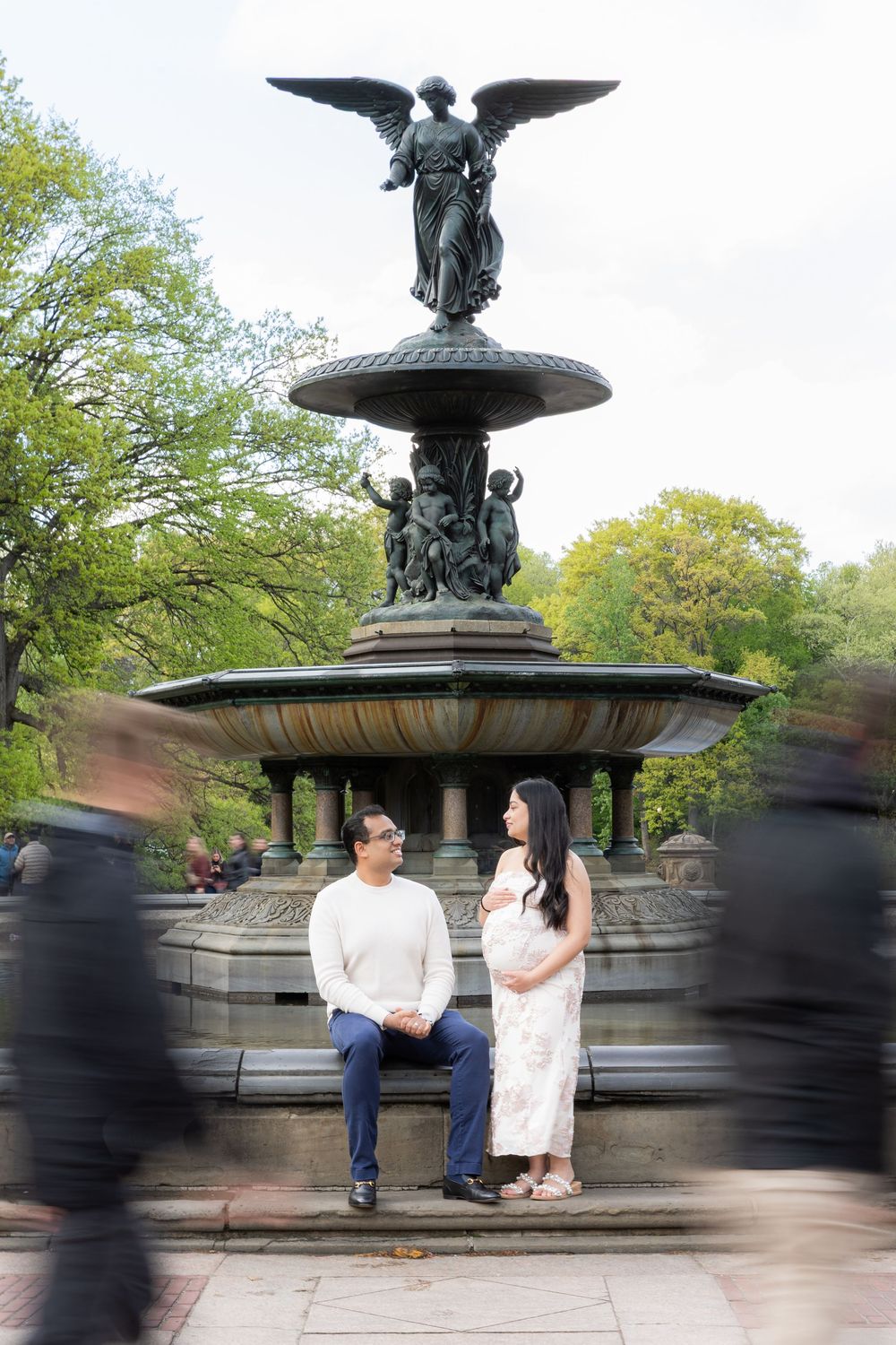 Couple sits at Bethesda Fountain in Central Park, NYC, with angel statue and blurred passersby.