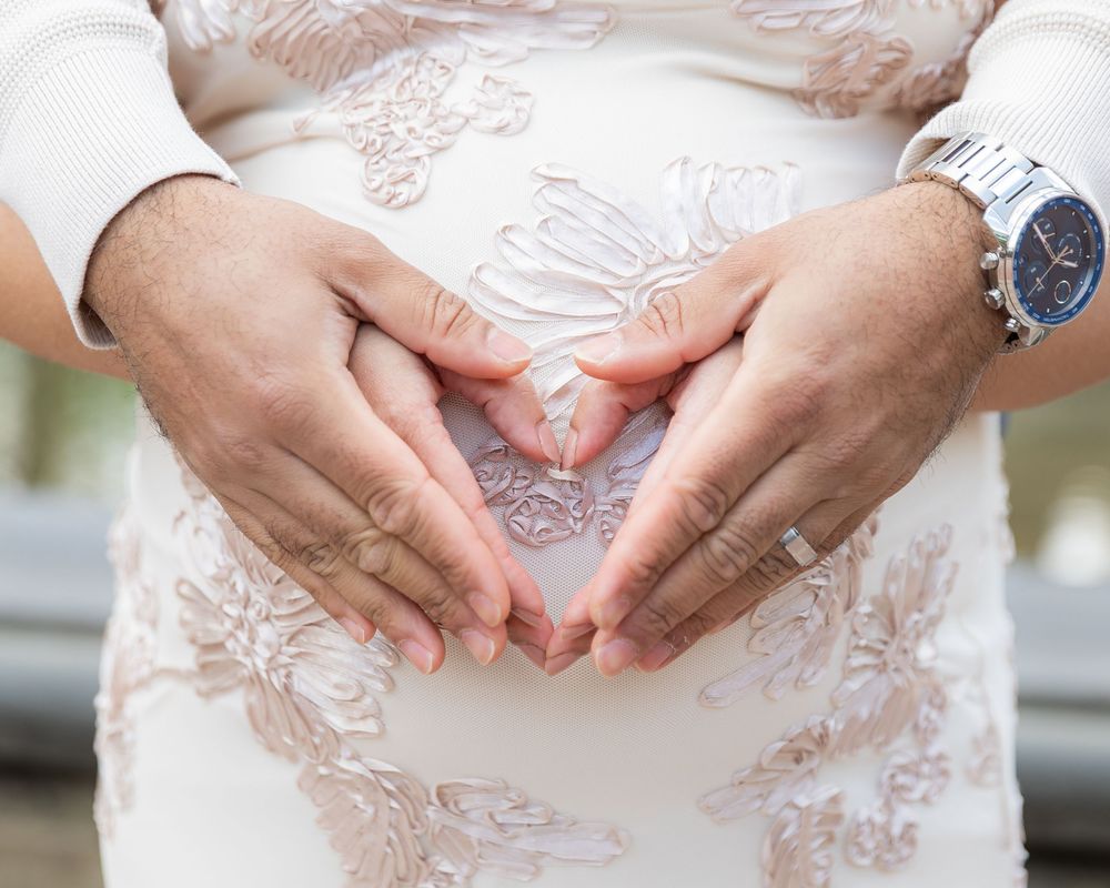 Couple forming a heart shape with hands on pregnant belly, wearing wedding rings and a silver watch.