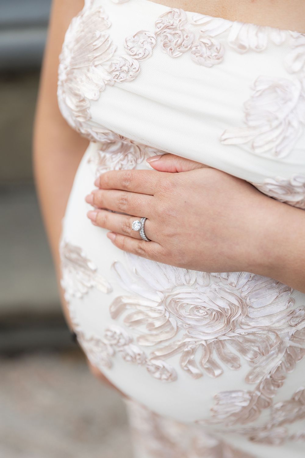 Pregnant woman in white floral lace maternity gown cradling baby bump, showcasing diamond engagement ring.
