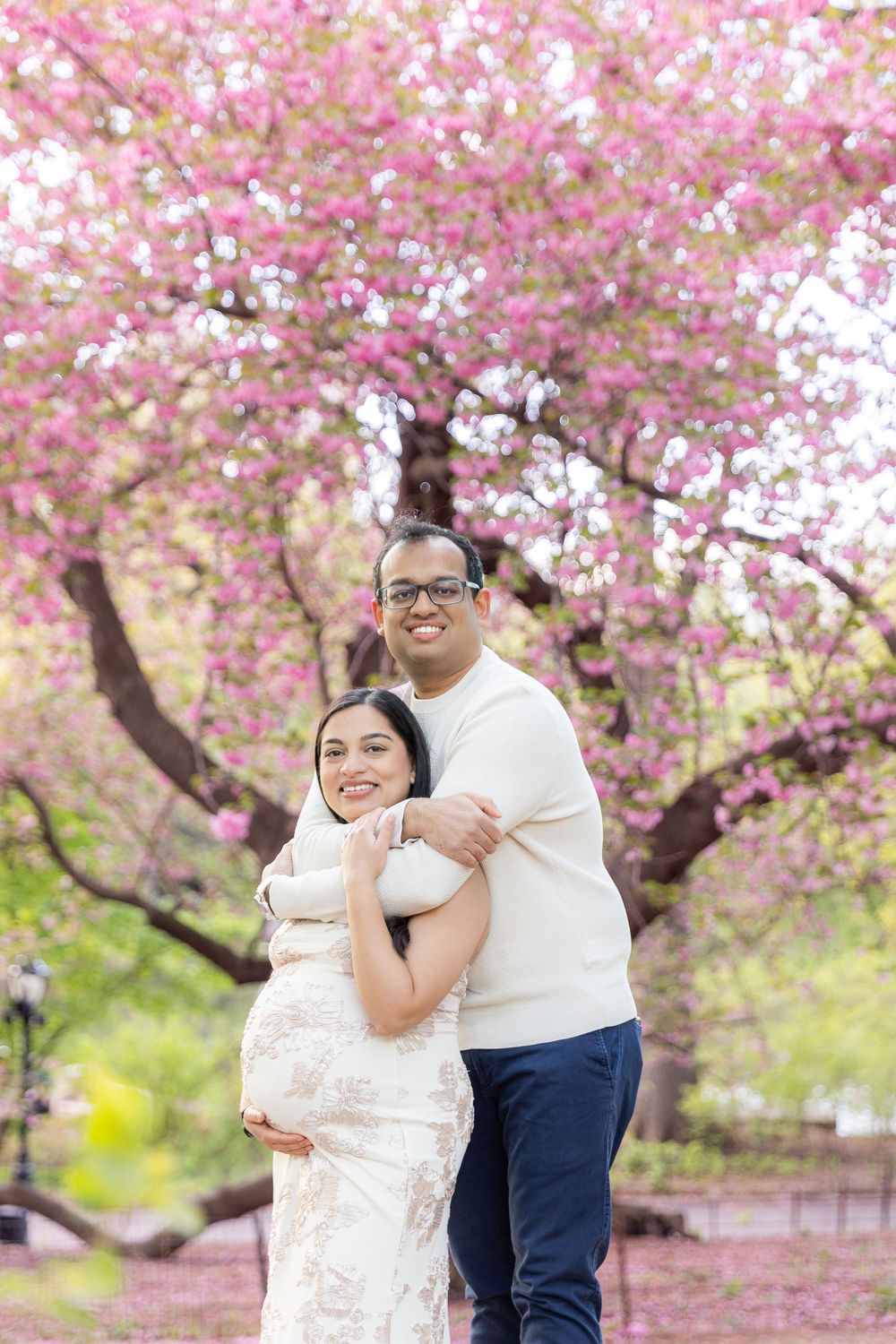 Couple posing in front of pink cherry blossom tree, pregnant woman in floral dress embraced by man in white sweater.