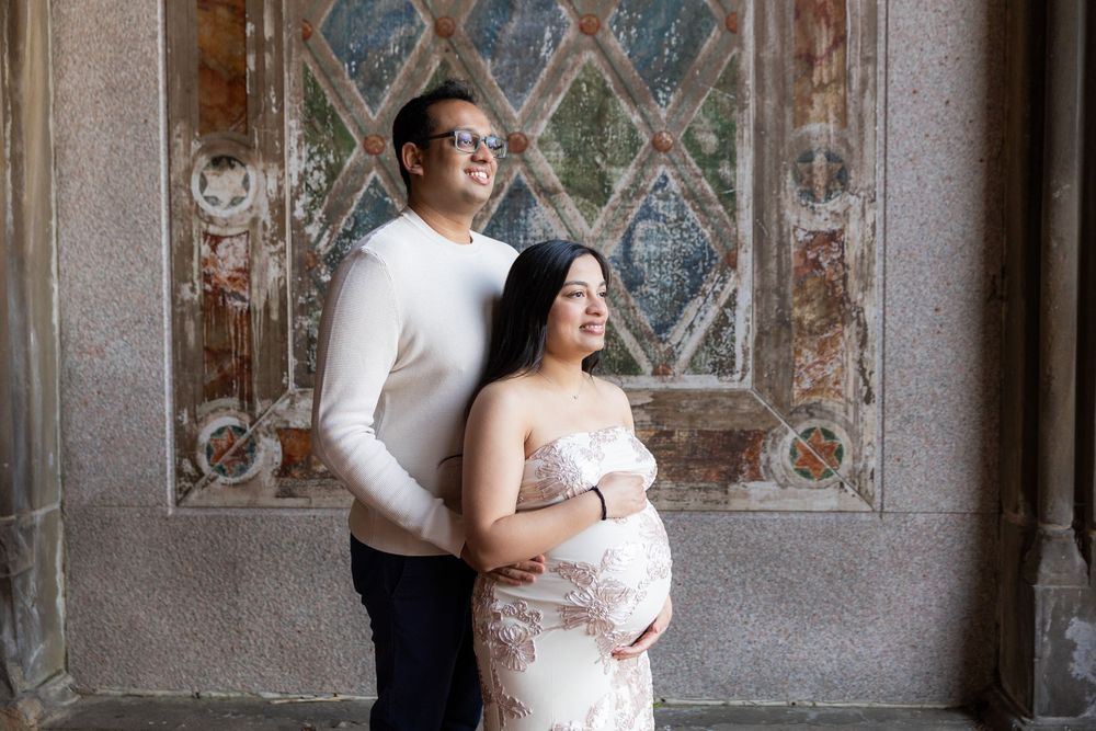 Couple poses for maternity photo in front of ornate mosaic wall, woman in lace gown cradling baby bump.
