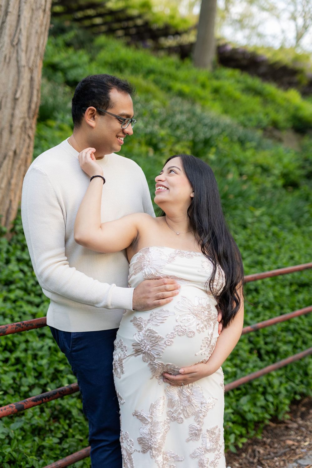 Happy expecting couple embracing outdoors, woman in floral lace maternity dress smiling at partner in green garden.