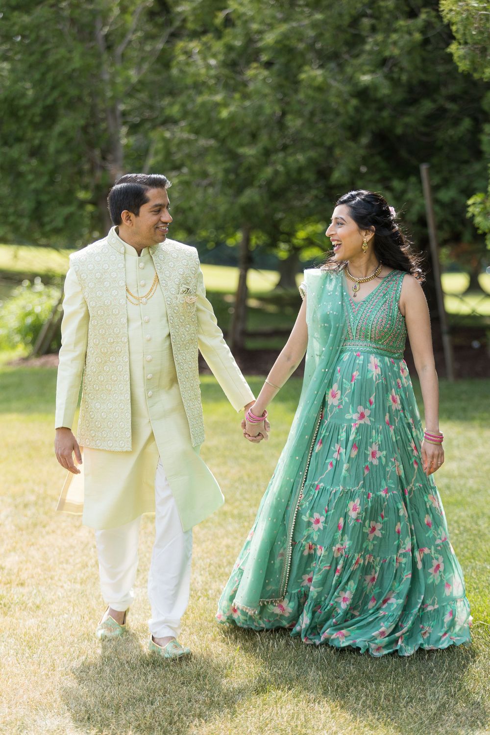 A couple in traditional South Asian attire walk hand in hand in a garden, wearing yellow and mint green formal wear.