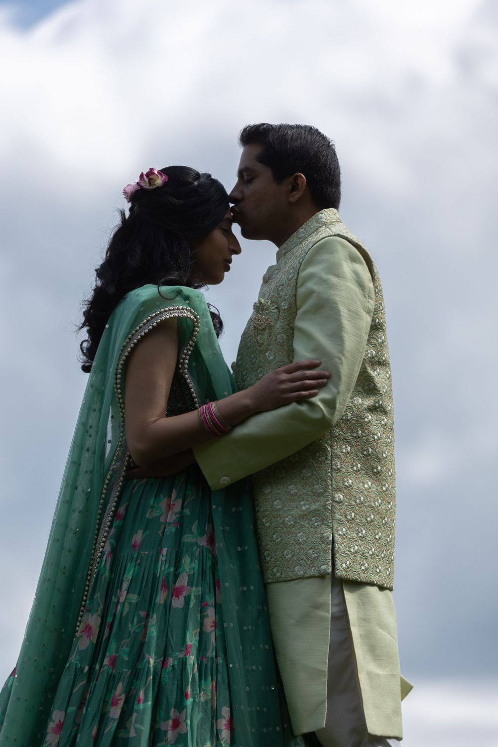 Couple in coordinating mint and cream outfits embrace against cloudy backdrop.