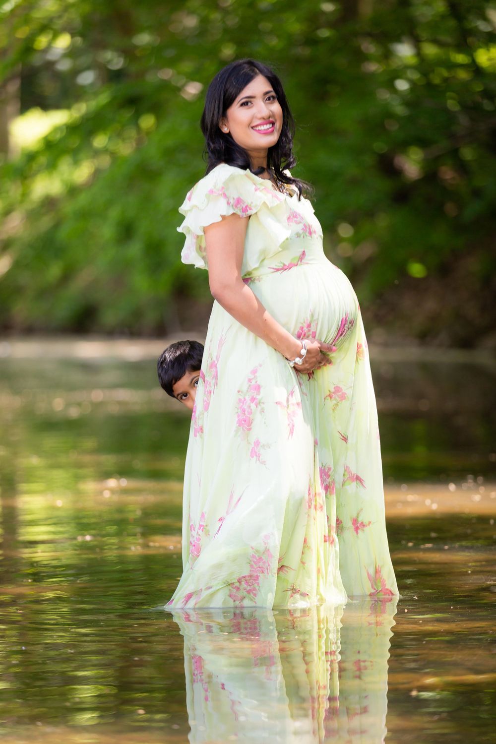 Expectant mother wearing a delicate floral dress stands in shallow water surrounded by greenery.