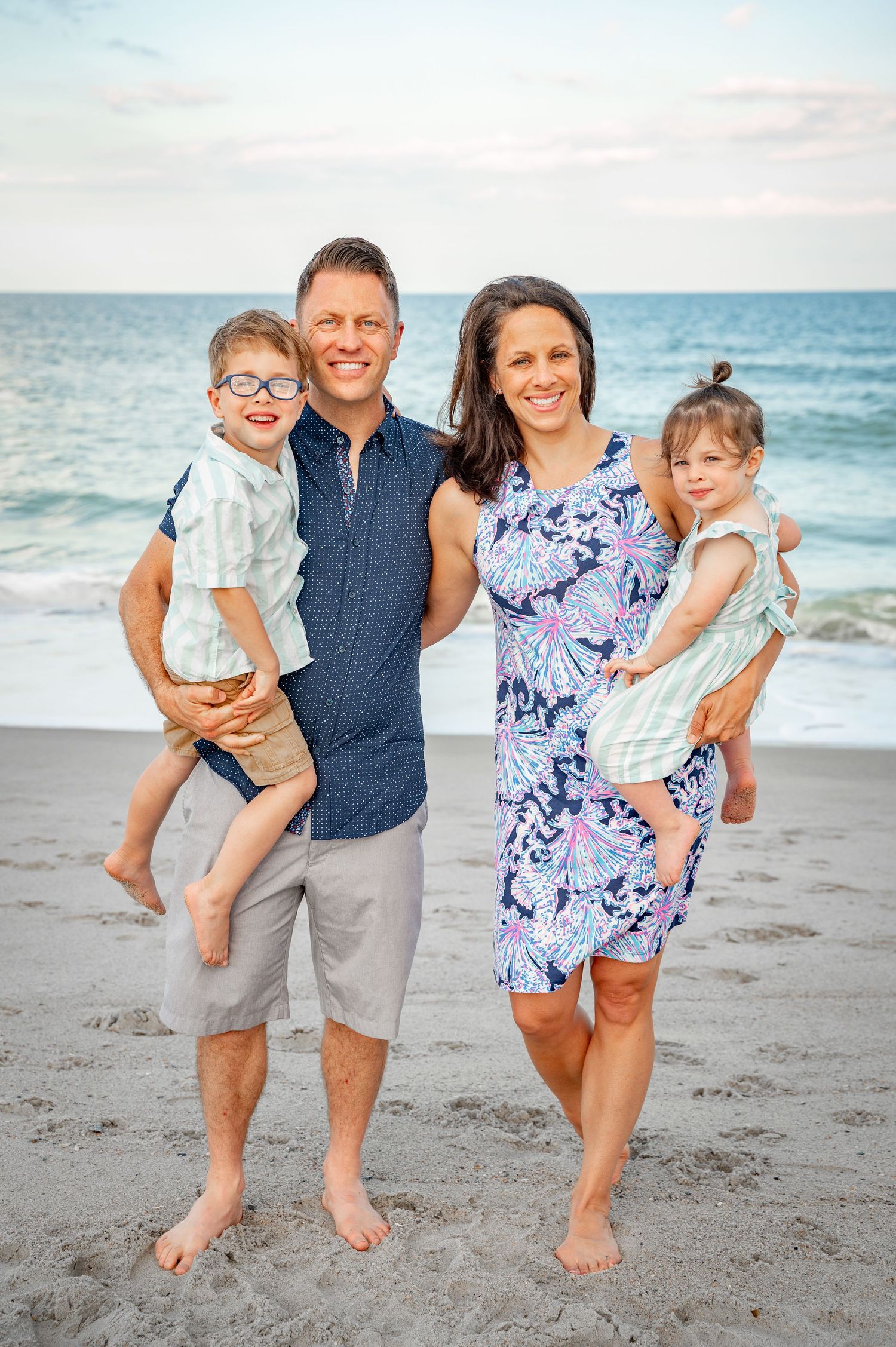 A happy family of four poses together barefoot on the beach at sunset.