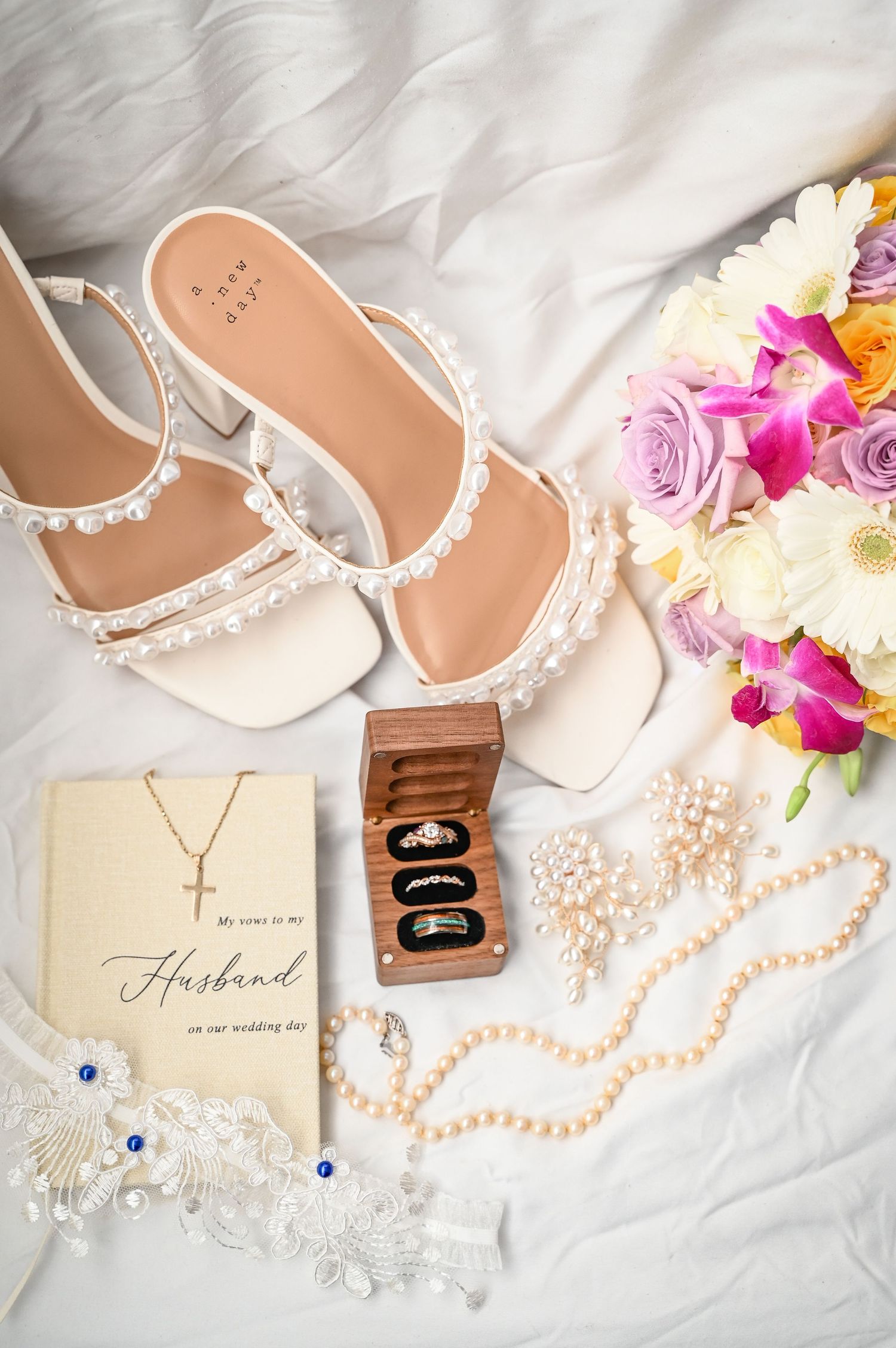 Wedding accessories including pearl-adorned sandals, floral bouquet, necklace, ring box and invitation card on white fabric.