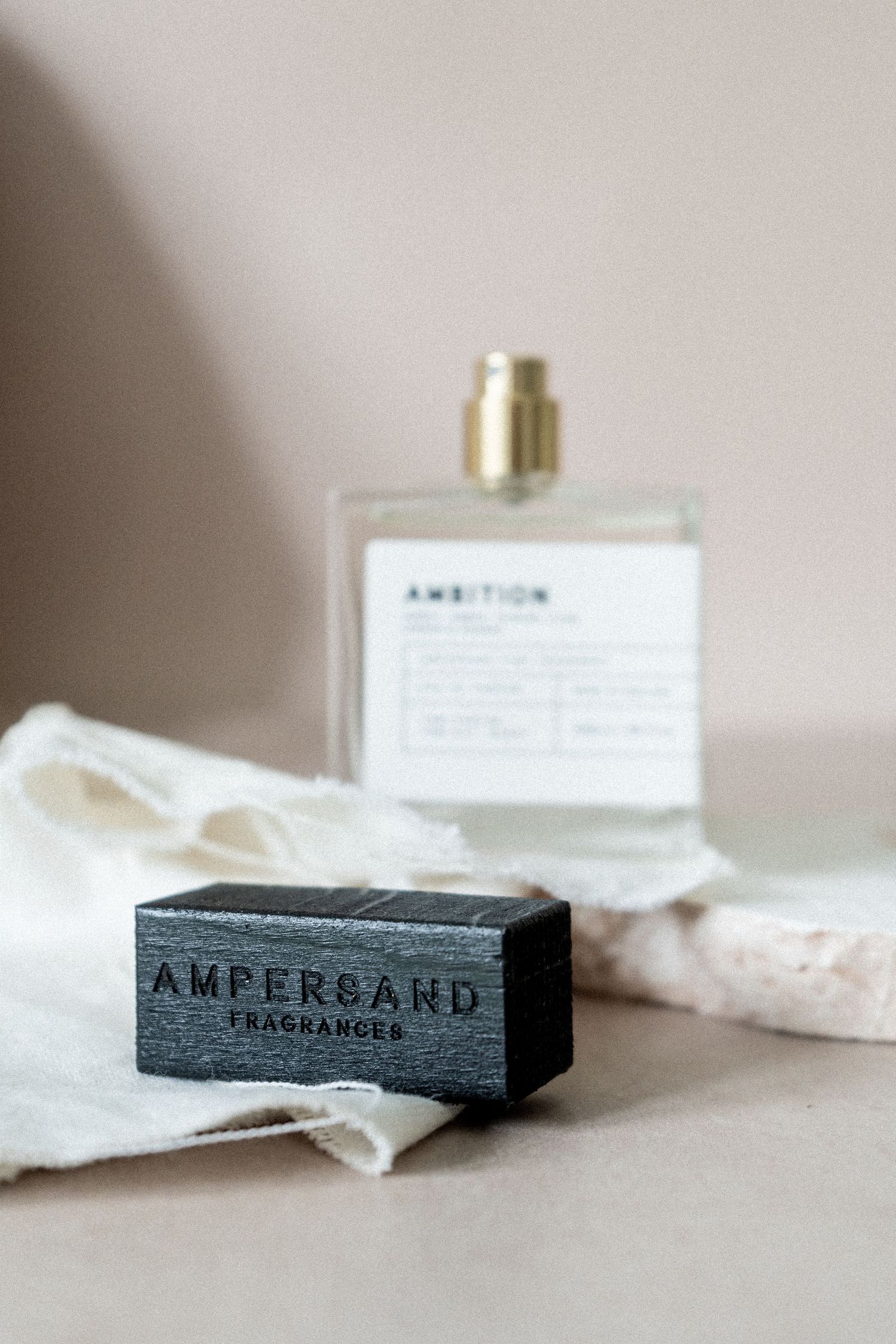 studio photo of perfume bottle with small textiles and a rock, sand coloured background