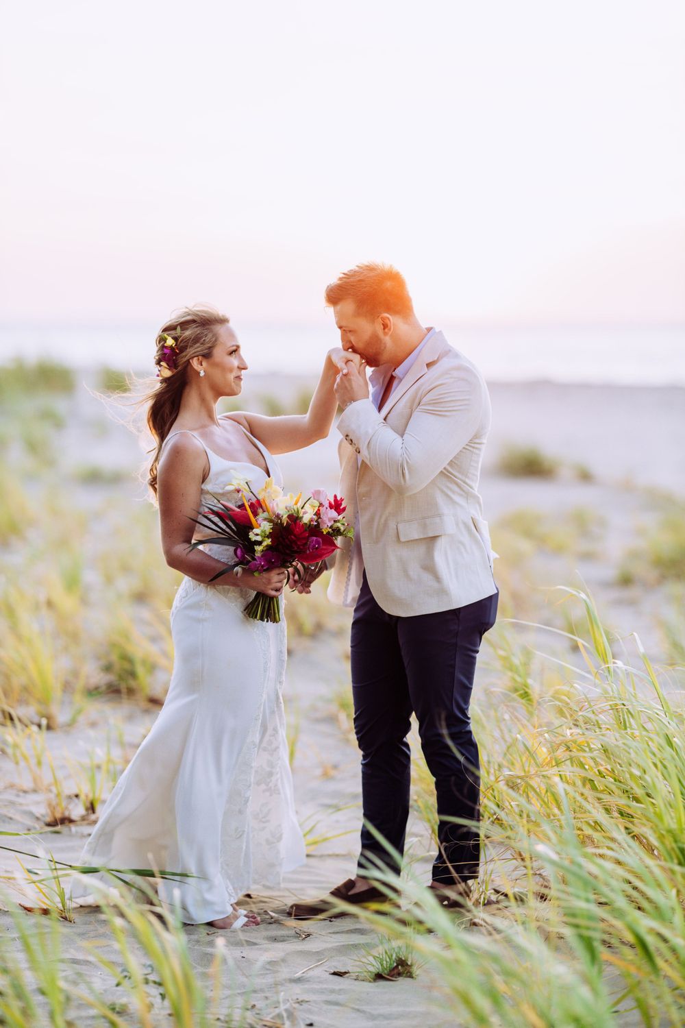 Sunset beach wedding portrait with couple holding tropical flowers among beach grass.
