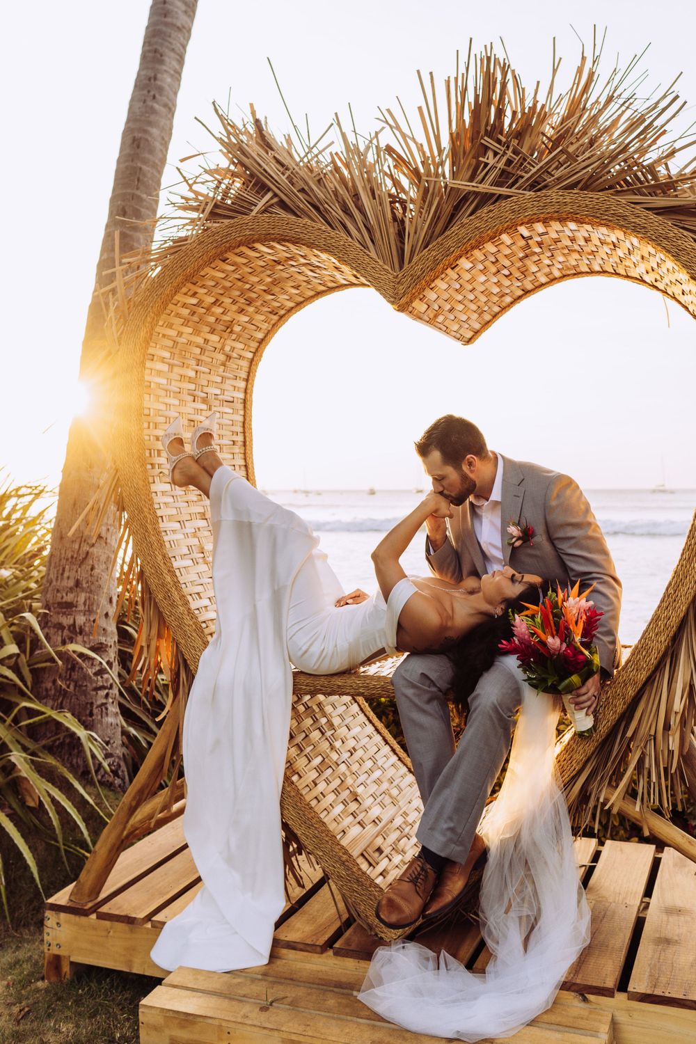 Romantic sunset moment at tropical beach wedding under woven heart-shaped arch.