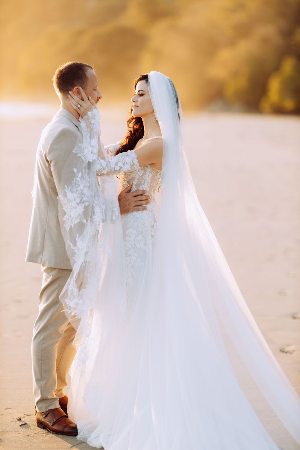 A romantic beach wedding photoshoot featuring a bride in a lace gown and groom in beige suit at sunset.