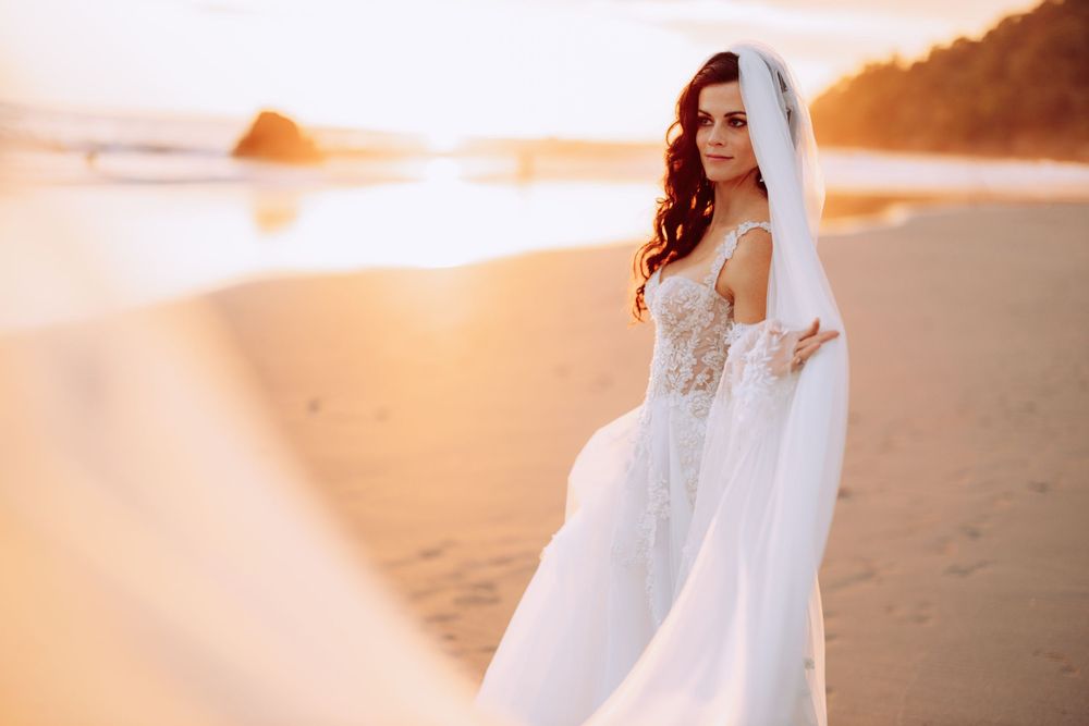 Bride poses on beach at sunset with dramatic veil and detailed lace gown.