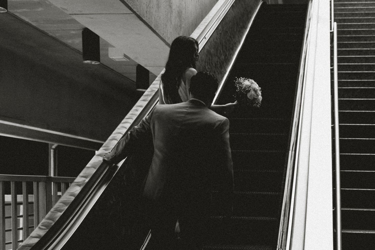 Black and white photo of a couple in their wedding outfits going up an escalator to catch the train with a moody vibe.