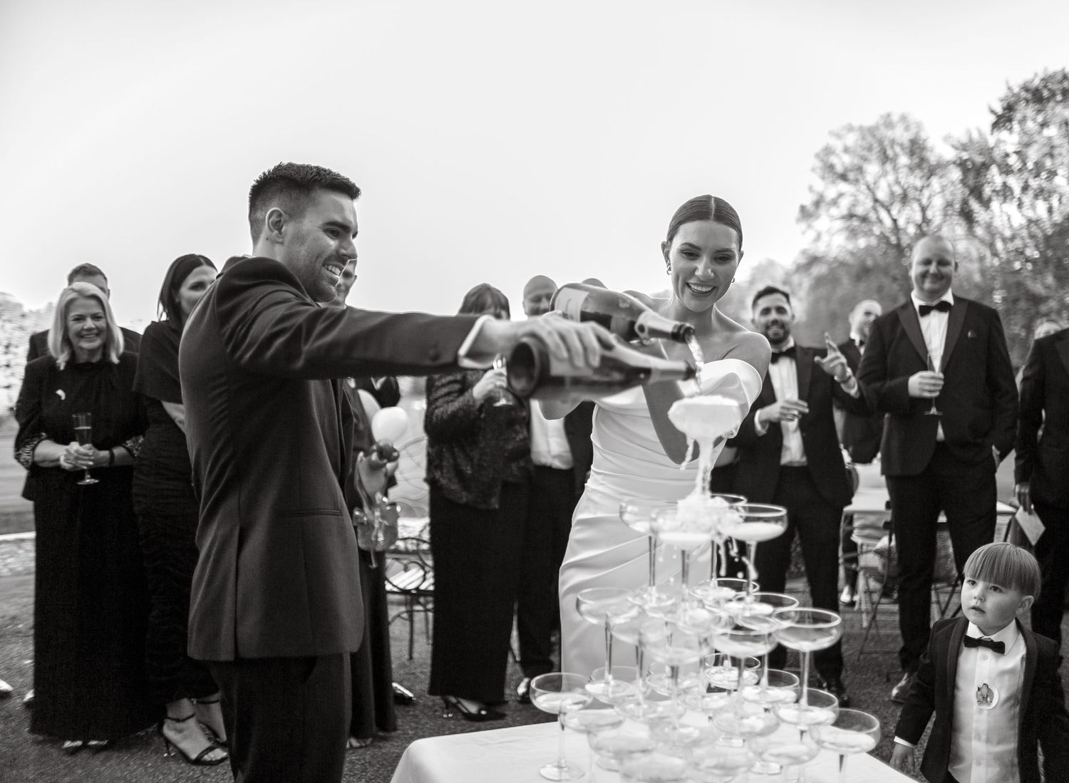Wedding guests pouring champagne into a tower of glasses at an outdoor celebration.