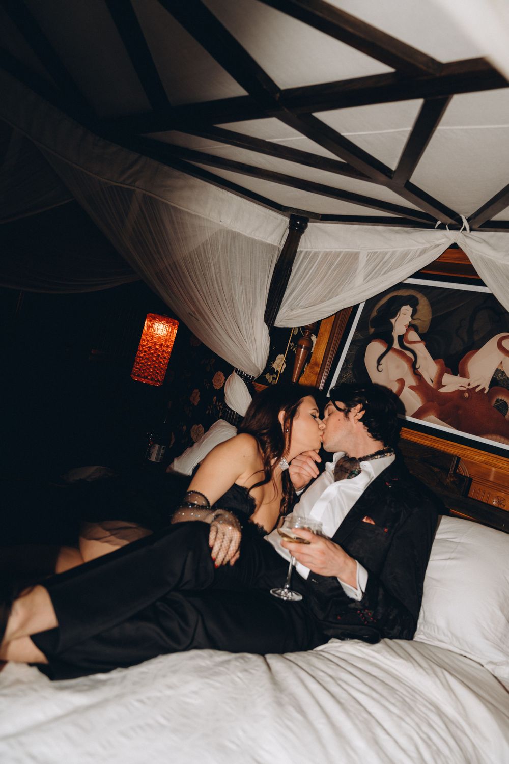 Romantic intimate couple lounging on white bed beneath draped canopy with orange lantern illuminating dark room.