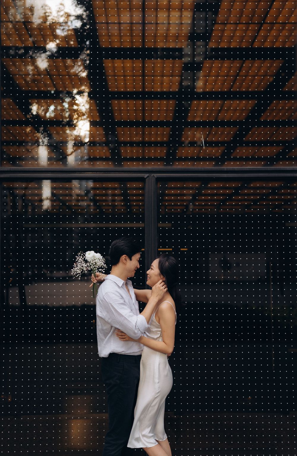 A romantic couple in formal attire embraces against a dark geometric background under warm lighting.