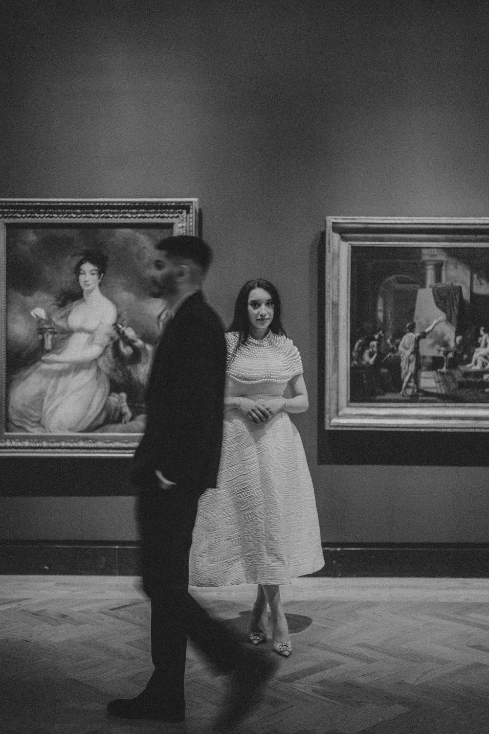 Museum visitors in formal attire view classical paintings in a gallery, captured in black and white.