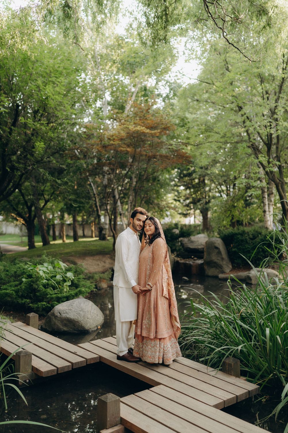 A couple in traditional South Asian attire stands together on a wooden bridge in a peaceful garden setting.